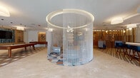This thoughtfully designed single-floor amenity space offers a dynamic blend of social, recreational, and relaxation areas unified by a cohesive contemporary aesthetic. The centerpiece of the space is a dramatic circular enclave defined by cascading chain-curtain walls suspended from a glowing ring fixture overhead, creating an intimate seating nook set atop a vibrant geometric patterned rug. Surrounding this focal point, the open-plan layout flows seamlessly between distinct activity zones. A game room features a billiards table alongside a mounted television framed in teal mosaic tile and set against warm wood paneling. A cozy lounge area is lined in natural wood paneling from floor to ceiling, complete with rich terracotta-toned sofas, built-in display shelving, and circular pendant lighting. The fully equipped kitchen boasts sleek white cabinetry with dark countertops, a bold geometric tile floor, built-in appliances, and ample counter space — all bathed in natural light from large windows. Additional seating nooks provide quieter retreat options, including a carpeted alcove with a wall-mounted TV. Light wood slatted partitions and curated wall art punctuate the space throughout, contributing to a warm, modern, and inviting atmosphere tailored for community gathering and recreation.