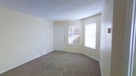 This single-floor space features a welcoming main living area finished with wall-to-wall carpeting in a neutral tan tone, providing a comfortable and practical foundation underfoot. The room is bathed in natural light, courtesy of two windows dressed with white horizontal blinds that offer views of the surrounding exterior. A classic six-panel white entry door is positioned between the windows, creating a clean and functional entryway focal point. The walls are painted in a soft, warm off-white hue that complements the light tones throughout the space. A textured popcorn ceiling adds a traditional finish overhead, and a smoke detector is visibly mounted for safety. Baseboards run along the perimeter of the room, and multiple electrical outlets are positioned conveniently along the walls. The open layout of the living area allows for versatile furniture arrangement. The space presents a straightforward, functional design that is move-in ready, making it well-suited for a variety of living arrangements. Overall, the room conveys a clean, modest, and practical ambiance throughout.