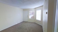 This single-floor space features a welcoming main living area finished with wall-to-wall carpeting in a neutral tan tone, providing a comfortable and practical foundation underfoot. The room is bathed in natural light, courtesy of two windows dressed with white horizontal blinds that offer views of the surrounding exterior. A classic six-panel white entry door is positioned between the windows, creating a clean and functional entryway focal point. The walls are painted in a soft, warm off-white hue that complements the light tones throughout the space. A textured popcorn ceiling adds a traditional finish overhead, and a smoke detector is visibly mounted for safety. Baseboards run along the perimeter of the room, and multiple electrical outlets are positioned conveniently along the walls. The open layout of the living area allows for versatile furniture arrangement. The space presents a straightforward, functional design that is move-in ready, making it well-suited for a variety of living arrangements. Overall, the room conveys a clean, modest, and practical ambiance throughout.