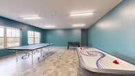 This game room in Madison, WI, offers a well-appointed recreational space designed for active social engagement. The single-floor layout provides a generous open area that comfortably accommodates multiple game stations simultaneously, including a foosball table, an air hockey table, and a full-size ping-pong table, allowing several activities to take place without crowding.

The room's design is defined by its bold teal-painted walls, which lend a vibrant, energetic character to the space. Durable stone-look tile flooring in warm, neutral tones runs throughout, providing a practical and attractive surface suited to high-traffic recreational use. Overhead fluorescent linear light fixtures ensure consistent, bright illumination across the entire room.

Natural light enters through multiple windows along one wall, offering views to the exterior and contributing to a lively, open atmosphere. High-top tables with bar stools are positioned near the windows, providing casual seating for spectators or those taking a break between games. A standard entry door provides access to the adjoining areas of the building. Overall, the space is clean, functional, and thoughtfully arranged to serve as an engaging recreational amenity.
