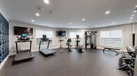 This fitness center is thoughtfully designed to support a comprehensive workout experience within a clean, modern environment. The space is laid out across a single floor and benefits from abundant natural light streaming through multiple large windows, offering views of the surrounding area and creating an open, energizing atmosphere.

The cardio zone is well-appointed with treadmills, stationary bikes, and elliptical trainers, each equipped with personal display screens. A wall-mounted flat-screen television adds to the workout experience. The flooring throughout is durable rubber sport tile in a speckled gray finish, providing both safety and comfort underfoot.

The strength training area features a multi-station cable machine, a flat utility bench, and a fully stocked dumbbell rack ranging across a wide variety of weights. A large mirror spans one wall, enhancing the sense of space and allowing users to monitor their form.

A standout design element is the bold navy blue wave-textured accent wall, which adds a striking visual contrast to the otherwise neutral gray palette of the room. Recessed lighting throughout ensures even, well-distributed illumination. The space also includes a defibrillator station, a wireless access point, and a keycard-secured entrance, reflecting a commitment to both safety and convenience.