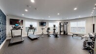 This fitness center is thoughtfully designed to support a comprehensive workout experience within a clean, modern environment. The space is laid out across a single floor and benefits from abundant natural light streaming through multiple large windows, offering views of the surrounding area and creating an open, energizing atmosphere.

The cardio zone is well-appointed with treadmills, stationary bikes, and elliptical trainers, each equipped with personal display screens. A wall-mounted flat-screen television adds to the workout experience. The flooring throughout is durable rubber sport tile in a speckled gray finish, providing both safety and comfort underfoot.

The strength training area features a multi-station cable machine, a flat utility bench, and a fully stocked dumbbell rack ranging across a wide variety of weights. A large mirror spans one wall, enhancing the sense of space and allowing users to monitor their form.

A standout design element is the bold navy blue wave-textured accent wall, which adds a striking visual contrast to the otherwise neutral gray palette of the room. Recessed lighting throughout ensures even, well-distributed illumination. The space also includes a defibrillator station, a wireless access point, and a keycard-secured entrance, reflecting a commitment to both safety and convenience.