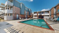 This outdoor communal space is centered around a generously sized swimming pool with brick coping, clearly marked depth indicators, and a no-diving designation — making it a safe and inviting retreat. The surrounding concrete pool deck is outfitted with lounge chairs, offering ample space for sunbathing beneath open skies. A standout feature is a vivid wave-themed mural painted on an exterior wall, adding a bold artistic touch to the environment.

The complex itself is composed of multi-story structures that blend classic brick facades with modern board-and-batten and corrugated metal siding in neutral tones of gray and blue. Upper-level balconies with white railings overlook the pool area, and a distinctive spiral staircase provides architectural interest while connecting levels near the gated entry points.

Adjacent to the pool, a covered pergola-style lounge area provides a shaded retreat furnished with seating including benches, chairs, and a coffee table — ideal for gathering out of direct sunlight. Brick columns support the overhead lattice structure, maintaining a cohesive aesthetic with the surrounding buildings. Iron fencing encloses the pool area, ensuring both security and an open, airy feel. Together, these amenities create a functional and visually engaging outdoor community hub.