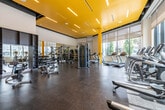 This contemporary fitness center is designed to inspire and energize, showcasing a striking yellow ceiling accented with sleek linear lighting that creates a vibrant, high-energy atmosphere throughout the main workout floor. The space is anchored by durable rubberized flooring and lined with floor-to-ceiling windows along one side, allowing abundant natural light to flood the interior while offering views of the surrounding urban environment.

The main gym floor is comprehensively outfitted with a wide selection of strength training equipment, including a multi-functional cable machine, a Smith machine with an adjustable bench, and a full rack of dumbbells spanning a broad weight range. The cardio zone features multiple treadmills, elliptical trainers, and stationary bikes, all positioned to take advantage of the natural light and city views.

A glass-enclosed studio provides a dedicated space for cycling classes, complete with premium spin bikes and warm wood flooring, separated from the main floor by full-height glass walls that maintain visual connectivity while offering an acoustically distinct environment. Mirrored walls throughout amplify the sense of space and provide functional feedback during workouts.

Additional amenities include accessory equipment storage, TRX suspension training anchors, and open floor areas for stretching and functional movement. The overall design blends bold architectural details with practical fitness functionality.