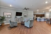 Located in Carrollton, Georgia, this single-floor community space is designed to foster comfort and social interaction in an open, welcoming environment. Rich hardwood flooring flows seamlessly throughout, anchoring each area with warmth and continuity.

The living area is thoughtfully arranged with plush upholstered seating, a wall-mounted flat-screen television, and a decorative credenza accented by framed artwork. Crown molding lines the ceilings, and a ceiling fan provides both style and function. Natural light filters in through large windows dressed with sheer curtains, enhancing the soft, sage-gray color palette that defines the space.

The kitchen is well-equipped with dark espresso cabinetry, light countertops, stainless steel appliances, and a generously sized island with bar seating — ideal for casual gatherings. Recessed lighting illuminates the workspace efficiently.

Multiple dining areas accommodate groups of varying sizes, each furnished with sturdy wooden tables and chairs. Botanical artwork and tasteful décor add character to these spaces. French doors in the dining area open to an outdoor area, inviting natural light and easy access to the exterior.

The overall ambiance is clean, cohesive, and purposefully designed for communal use, blending functional layout with a refined, residential aesthetic throughout.