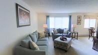 This single-floor space presents a comfortable and thoughtfully arranged living area that emphasizes a calm, cohesive aesthetic. The sitting room serves as the focal point, furnished with matching soft blue upholstered sofas and an accent armchair, all centered around a patterned ottoman that doubles as a coffee table. A tray with glassware rests atop the ottoman, adding a functional decorative touch.

Natural light enters through a large front-facing window dressed with vertical blinds and complementary blue curtain panels, lending the room a bright yet relaxed atmosphere. Neutral beige carpeting runs throughout, contributing warmth underfoot and reinforcing the understated color scheme. The textured ceiling adds subtle visual interest overhead.

Flanking table lamps on either side of the seating arrangement provide balanced ambient lighting, while a framed floral artwork on the left wall introduces a gentle pop of color. The overall layout is practical and open, with a clear sense of flow and purpose. The space is well-suited for relaxed everyday living, offering a welcoming environment that balances comfort with a tidy, coordinated interior style.