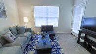 This single-floor space centers around a well-appointed living room that balances functionality with a clean, contemporary aesthetic. The room features neutral-toned walls and light beige wall-to-wall carpeting, creating a warm and inviting foundation. Natural light filters through a large window fitted with white horizontal blinds, while an additional window or glass door to the right further brightens the space.

The living area is thoughtfully arranged with a grey upholstered sofa, a sleek black tufted accent chair with a matching ottoman, and a dark rectangular coffee table. A bold blue and white abstract-patterned area rug anchors the seating arrangement, adding a striking visual focal point. Decorative accents, including a dark glass vase and a blue orb sculpture, are displayed on the coffee table, contributing to the room's cohesive color palette. A floor lamp in the corner provides supplemental ambient lighting. White baseboards trim the perimeter of the room, offering a crisp contrast to the neutral walls. The overall ambiance is modern and tidy, with a coordinated design scheme that feels both livable and visually engaging.