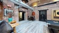 This single-floor clubhouse showcases a distinctive industrial-chic character defined by exposed brick walls, visible ductwork, and dark structural elements throughout. The space is generously sized, offering multiple distinct zones that cater to a variety of social activities. A lounge area near the entrance features rich leather seating, including a deep black leather sofa and red leather armchairs arranged around a rustic wood coffee table, with framed black-and-white photography and a large mirror adorning the brick walls. Adjacent to this seating area, a wall-mounted flat-screen television creates an ideal media viewing space. The main open area is anchored by a bar-height counter lined with black wooden stools, accompanied by a vending machine and beverage station, making it well-suited for casual gatherings. Dark tile flooring transitions to light wood-style planking across different zones, adding visual interest and helping to define each area. A black metal staircase provides architectural focus, while pool cues and game signage hint at additional recreational amenities. The combination of raw brick, high ceilings, industrial fixtures, and warm, comfortable furnishings creates a relaxed yet stylish communal atmosphere.
