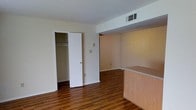 This single-floor space showcases a practical and open layout designed for comfortable everyday living. The flooring throughout is a rich, dark hardwood-style material that adds warmth and continuity across the main living area. A notable architectural element is the partial dividing counter constructed from light wood with a laminate top, which subtly separates functional zones within the space while maintaining an open feel.

A built-in closet with a standard rod for hanging storage is conveniently positioned along one wall, offering accessible organization. The adjacent open passageway leads into a secondary area featuring a textured, warm-toned accent wall, adding a subtle visual contrast to the otherwise neutral cream-colored walls throughout.

Overhead, a ceiling-mounted HVAC vent ensures climate control, and the smooth white ceilings contribute to a clean, uncluttered aesthetic. The space is well-lit with an even, neutral light that highlights the contrast between the dark flooring and light-colored walls. Overall, the layout is straightforward and functional, providing a versatile foundation well-suited for a variety of living arrangements.