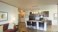 This single-floor residence offers a thoughtfully designed open-concept layout that seamlessly connects the kitchen, dining, and living areas into one cohesive and functional space. The kitchen is a standout feature, showcasing rich dark espresso cabinetry, granite countertops, and black appliances including a refrigerator, range, and built-in microwave. A breakfast bar with seating for three leather-upholstered bar stools serves as both a casual dining spot and a natural dividing element between the kitchen and living space. Pendant lighting fixtures hang above the bar, complemented by a track lighting system overhead that illuminates the kitchen workspace effectively. The living area extends naturally from the kitchen, anchored by warm hardwood-style flooring that runs throughout the space, contributing to a cohesive and inviting aesthetic. Neutral-toned walls keep the interior bright and versatile, while tasteful artwork adds personality and warmth. A hallway visible from the main living area leads to additional rooms, suggesting a well-organized floor plan. The overall ambiance is modern yet welcoming, blending practical design with stylish finishes that make this a comfortable and visually appealing space.