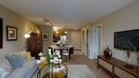 This single-floor residence showcases an open-concept design that integrates the living, dining, and kitchen areas into one cohesive, functional space. The living area is anchored by a sleek glass coffee table and a comfortable sofa, complemented by a dark wood display cabinet that adds both storage and visual interest. Warm neutral tones on the walls are enhanced by recessed lighting and a track lighting fixture overhead, creating a bright and inviting atmosphere throughout.

The dining area features a round dark-toned table surrounded by contemporary chairs, positioned beneath elegant pendant lighting that draws the eye upward. A light-colored area rug grounds the space and adds texture underfoot. Adjacent to the dining area, the kitchen is equipped with stainless steel appliances, warm wood cabinetry, and a pendant light above, offering both practicality and style.

Hardwood-style flooring extends throughout the main living areas, contributing to a clean, continuous aesthetic. A hallway visible in the background leads to additional rooms, hinting at further private spaces within the residence. The overall ambiance is modern yet warm, making it an approachable and functional home.
