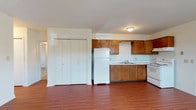 This single-floor unit offers a straightforward, functional layout that distributes living, sleeping, and kitchen spaces efficiently across one level. The open living area is finished with rich, wood-look laminate flooring that runs throughout the main living and kitchen zones, giving the space a warm and cohesive feel. Two entry points with exterior doors provide convenient access, and baseboard heating units are present throughout, ensuring consistent warmth.

The kitchen features warm-toned wood cabinetry on both upper and lower sections, providing ample storage. White appliances include an electric coil range, a refrigerator, and an under-cabinet range hood. A double stainless steel sink with a window-facing position adds practicality to the workspace. A large closet adjacent to the kitchen area offers additional storage.

Multiple bedrooms are finished with neutral wall-to-wall carpeting and individual windows that allow natural light to filter in with views of greenery. Each room includes baseboard heating and presents a clean, versatile space. The bathroom is appointed with a full tub and shower combination, featuring a distinctive light blue tub contrasted by white subway tile walls and a window above, lending a classic, retro character to the space.