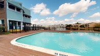 This outdoor amenity space centers around a large, rectangular swimming pool with clear turquoise water, surrounded by a broad wood-plank deck that provides ample room for poolside seating and leisure. The pool area is enclosed by black iron fencing, offering both security and an open, airy feel. A gated entry point leads to the deck, which is accented by colorful flowering plants near the entrance of an adjacent light blue-sided building that houses supporting facilities.

The deck extends toward a waterfront boundary, where a second iron fence separates the pool area from a scenic lake or pond beyond. Views across the water reveal a neighboring residential community, adding a pleasant backdrop to the outdoor setting. Seating areas on the deck allow residents to enjoy the open-sky atmosphere and waterfront scenery.

The surrounding grounds include mature trees, well-maintained shrubs, and green lawns that frame the property with natural landscaping. The brick and blue-sided residential buildings nearby reflect a mixed architectural character typical of established communities. It is worth noting that portions of the wood decking show visible wear and structural deterioration, including areas with significant damage, indicating a need for maintenance or repair. Overall, the space offers a functional and scenic outdoor retreat with strong waterfront appeal.