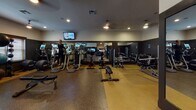 This fitness center in State College, Pennsylvania offers a comprehensive workout environment designed to support a wide range of exercise routines. The space is finished with durable, shock-absorbing rubber flooring in a warm tan tone, providing a safe and practical surface throughout. Dark accent walls contrast with lighter upper walls and a white ceiling, creating a modern, energetic atmosphere enhanced by recessed lighting and ceiling fans that ensure comfort during workouts.

The cardio area features multiple treadmills and elliptical machines, each equipped with individual digital displays, allowing users to track their performance. Mounted flat-screen televisions are positioned throughout the space, keeping exercisers entertained during their sessions. A row of windows fitted with wood-toned blinds brings in natural light while maintaining privacy.

The strength training section is well-appointed with a cable machine and multi-function weight station, a dumbbell rack stocked with a range of free weights, and an adjustable bench. Large wall mirrors expand the sense of space and allow users to monitor their form. A water dispenser is conveniently placed within reach. The layout is thoughtfully organized to allow multiple users to exercise simultaneously without congestion, making this fitness center a functional and inviting amenity.