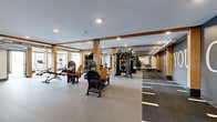 Located at 4066 Lexington Ave N in Shoreview, Minnesota, this professionally designed fitness facility spans a generously sized single floor, offering a comprehensive workout environment across multiple distinct zones. The main exercise area is defined by warm wood beam columns and recessed lighting, creating an inviting yet energetic atmosphere. It is outfitted with a wide array of strength training equipment, including multi-functional cable machines, power racks, barbells, weight plates, and an extensive dumbbell rack, all set on durable rubberized flooring. Motivational signage — including a bold "YES. YOU. CAN." mural — adorns the dark accent walls, reinforcing an encouraging training environment. Large mirrors throughout enhance spatial depth and support proper form during workouts. A dedicated cardio zone features treadmills, an elliptical, and a rowing machine, bathed in abundant natural light from floor-to-ceiling windows that offer outdoor views. A separate room serves as an indoor golf simulator space, complete with green turf flooring, a projector, and large windows that flood the area with natural light. The facility also includes additional support spaces, completing a well-rounded, amenity-rich fitness destination.