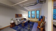 This single-floor urban loft-style unit blends industrial design with modern comfort, highlighted by exposed silver ductwork running along the ceilings — a distinctive architectural detail that defines the character of the space. The living area is generously proportioned, featuring large city-facing windows that frame striking elevated views of the surrounding skyline. Large-format tile flooring, track lighting, and clean neutral walls complete the contemporary aesthetic. A bold blue overdyed area rug and tufted gray sofa anchor the living space, complemented by a warm wood media console and wall-mounted art panel.

The kitchen is well-appointed with warm-toned wood cabinetry, granite countertops, stainless steel appliances, and a central island with bar seating for three — lit dramatically by two oversized pendant drum lights. Ample cabinetry and full-height storage make it highly functional.

The bedroom offers a calm retreat with plush carpeting, dual windows overlooking the cityscape, and room for a full-sized bed with nightstands. The full bathroom features a tiled shower-tub combo, clean white fixtures, and a wood-accented vanity with a white countertop, providing a crisp, functional finish throughout.