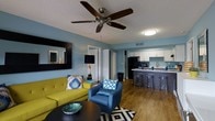 This single-floor space offers an inviting open-concept layout that seamlessly connects the living area and kitchen, creating a cohesive and functional environment. The living room is anchored by rich hardwood-style flooring and features soft blue-toned walls that give the space a calm yet energetic feel. A ceiling fan with wood-finished blades serves as a practical overhead focal point, while a bold chartreuse sofa and a vibrant blue accent chair introduce lively pops of color. A geometric black-and-white area rug grounds the seating arrangement, complemented by a sleek round coffee table. The kitchen is defined by crisp white cabinetry, a dark countertop peninsula with acrylic bar stools, and a full suite of black appliances, including a refrigerator. The peninsula provides additional seating and serves as a natural divider between the living and cooking spaces. Throughout, the design reflects a modern aesthetic with carefully coordinated furnishings and finishes. A mirrored wall accent in the living area adds depth and dimension to the space. Interior doors lead to additional areas, completing this well-appointed, stylishly designed single-level residence.