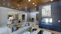 This single-floor residence showcases a thoughtfully designed open-concept layout that seamlessly connects multiple living areas. The living room serves as the centerpiece, highlighted by a dramatic wood-planked ceiling fitted with an array of recessed lights that cast a warm, ambient glow throughout the space. Light hardwood flooring runs continuously across the open floor plan, enhancing the sense of flow and continuity.

A bold, dark-framed double-sided fireplace anchors the living area, topped with an oversized abstract artwork that adds a striking visual element. The living space is furnished with a generously sized neutral-toned sofa accented with patterned and solid blue throw pillows, complemented by a marble-topped coffee table.

Beyond the main living area, a dining zone is visible, featuring bar-height seating and additional lounge-style furniture, suggesting a flexible entertaining layout. A secondary lounge area is partially visible through an open passage, indicating the expansive, multi-zone design of the interior. The overall ambiance is modern and refined, balancing warm natural textures with a cool, neutral color palette.