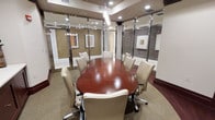 This single-floor conference room offers a polished and professional environment designed for productive meetings and collaborative sessions. The centerpiece is a large, oval-shaped table finished in rich mahogany, surrounded by cream-colored leather executive chairs with caster bases, comfortably accommodating up to ten participants.

The room is enclosed by floor-to-ceiling glass partition walls framed in sleek metallic accents, providing both transparency and a sense of openness while maintaining acoustic separation from surrounding spaces. The glass panels are fitted with sliding doors, allowing flexible access.

Overhead, a combination of recessed ceiling lights and elegant hanging pendant fixtures provides well-distributed, warm illumination that enhances the room's refined atmosphere. The neutral-toned carpet and warm wall finishes contribute to a sophisticated yet welcoming ambiance.

Framed artwork on the walls adds a subtle decorative touch without distracting from the room's professional character. The overall design balances functionality with a corporate aesthetic, making it well-suited for executive meetings, client presentations, and team discussions.