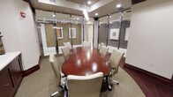 This single-floor conference room offers a polished and professional environment designed for productive meetings and collaborative sessions. The centerpiece is a large, oval-shaped table finished in rich mahogany, surrounded by cream-colored leather executive chairs with caster bases, comfortably accommodating up to ten participants.

The room is enclosed by floor-to-ceiling glass partition walls framed in sleek metallic accents, providing both transparency and a sense of openness while maintaining acoustic separation from surrounding spaces. The glass panels are fitted with sliding doors, allowing flexible access.

Overhead, a combination of recessed ceiling lights and elegant hanging pendant fixtures provides well-distributed, warm illumination that enhances the room's refined atmosphere. The neutral-toned carpet and warm wall finishes contribute to a sophisticated yet welcoming ambiance.

Framed artwork on the walls adds a subtle decorative touch without distracting from the room's professional character. The overall design balances functionality with a corporate aesthetic, making it well-suited for executive meetings, client presentations, and team discussions.