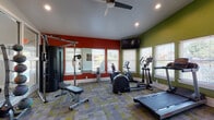 This fitness center is designed to support a full range of workout routines in a dynamic, motivating environment. The space is characterized by bold accent walls in deep red and olive green, which contrast with the neutral gray tones of the textured, patterned carpet tiles underfoot. Recessed lighting and a ceiling fan ensure the space remains well-lit and comfortable throughout any workout session.

The cardio zone features multiple treadmills, elliptical trainers, and a stationary bike, all positioned along the windowed walls to allow natural light to flow in and provide views of the surrounding outdoors. A wall-mounted flat-screen television is positioned prominently for entertainment during workouts. Large mirrors along one wall enhance the sense of space and allow for form-checking during exercises.

The strength training area is comprehensively stocked with a full rack of dumbbells in varying weights, medicine balls, stability balls, and a multi-function cable machine with an adjustable bench. High ceilings, particularly notable in the free weights section, contribute to an open, airy feel. Large interior-facing glass panel windows connect the fitness center visually to an adjacent lounge area. Fitness-themed artwork adds personality and energy to the walls, completing the motivating atmosphere of this thoughtfully designed facility.