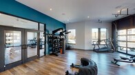 This contemporary fitness center is designed to accommodate a wide range of workout styles within a single, open-plan layout. The space is finished with durable wood-look flooring throughout and features crisp white walls accented by bold teal and dark charcoal feature walls, creating a dynamic and motivating atmosphere. Recessed lighting illuminates the interior, while large windows and expansive floor-to-ceiling glass garage doors flood the space with natural light and provide views of the surrounding exterior.

The facility is divided into distinct functional zones. A dedicated strength and functional training area is anchored by a fully stocked multi-tiered rack holding kettlebells, medicine balls, and slam balls, complemented by a heavy punching bag and a large training tire for high-intensity workouts. Full-length mirrors are mounted on the teal accent wall and near the glass-paneled entry doors, supporting form monitoring and making the space feel larger.

A separate cardio zone houses treadmills and an air resistance bike, while an additional area features two interactive cycling bikes positioned alongside a modular shelving unit. The glass garage doors open the fitness center to the outdoors, offering a flexible indoor-outdoor training experience. The overall design balances industrial-modern aesthetics with practical, performance-focused amenities.