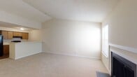 This single-story residence offers a practical and comfortable layout with distinct living, kitchen, and bedroom areas. The spacious living room is a standout feature, showcasing a classic fireplace flanked by two windows that allow natural light to fill the room. Neutral-toned carpet and vaulted ceiling details lend the space an open, airy feel.

The kitchen is designed for functionality, featuring warm wood-toned cabinetry, black appliances including a range and refrigerator, light countertops, and a peninsula with a built-in sink that opens toward the living area. Wood-look flooring grounds the kitchen space and provides a clean contrast to the carpeted areas.

The bedroom is generously sized with a large window providing ample natural light and an adjoining walk-in closet offering notable storage. An additional carpeted room with a built-in ledge and vaulted ceiling could serve as a secondary bedroom, office, or flex space.

The bathroom includes wood-look flooring, a wood-toned vanity, open shelving, a large mirror, and a grab bar, combining practicality with a clean, neutral aesthetic. Throughout the space, textured ceilings and white baseboards provide consistent architectural detailing.