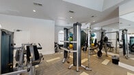 Located at 8787 Woodway Dr in Houston, Texas, this fitness center offers a well-appointed workout facility designed to support a comprehensive exercise routine. The single-floor space is thoughtfully laid out to maximize functionality while maintaining an open, airy feel. Neutral-toned, patterned carpet tiles cover the floor throughout, lending a clean and modern aesthetic to the room.

The strength training area is equipped with a variety of cable machines, including Magnum and Matrix branded units, along with a power rack, weight bench, and an assortment of medicine balls and dumbbells. Full-length mirrored walls amplify the sense of space and allow users to monitor their form during workouts.

The cardio section lines the perimeter with a row of elliptical trainers, treadmills, and a recumbent bike, several of which feature personal monitors. Wall-mounted flat-screen televisions are positioned throughout for entertainment during workouts. Recessed lighting ensures the interior is well-lit, while arched and large rectangular windows invite natural light and offer views of the surrounding outdoor amenity areas, including glimpses of a pool and covered patio. The elevated ceilings with clerestory-style skylights further enhance the bright and energizing atmosphere of the space.
