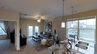 This single-floor space showcases an open-concept design that integrates the kitchen and living area into one cohesive, flowing layout. The living area is comfortably sized, anchored by plush carpeting and centered around a gray sofa and coordinating accent chairs arranged on a decorative area rug. A ceiling fan with a light fixture provides both airflow and ambient lighting overhead, while pendant lights hang above the kitchen counter, adding a warm, modern touch.

The kitchen features granite countertops and a sink positioned beneath a large window, allowing natural light to pour in. The open counter space transitions smoothly into the living area, making it ideal for entertaining.

Large windows on multiple walls flood the interior with natural light and offer views of the surrounding exterior. French doors lead directly to a private balcony, further enhancing the connection between indoor and outdoor living. Decorative wall art featuring framed feather prints adds a stylish, cohesive aesthetic. Overall, the space presents a warm, well-lit, and thoughtfully arranged interior with a contemporary residential feel.