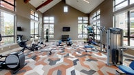 This fitness center is a thoughtfully designed workout space that combines striking architectural details with a comprehensive array of exercise equipment. The room commands attention with its soaring vaulted ceiling, accented by exposed wooden beams and clerestory windows that flood the space with natural light from multiple directions. Large floor-to-ceiling windows line the walls, offering open views of the surrounding landscape and lush greenery, creating an energizing and airy atmosphere throughout.

The flooring makes a bold design statement, featuring a vivid geometric pattern in terracotta, gray, and white tones that adds visual energy to the space. Cardio equipment, including treadmills and elliptical trainers, is positioned along the windowed walls to allow users to enjoy the outdoor views during their workouts. The strength-training area is well-stocked with selectorized cable machines, weight benches, and a full dumbbell rack, catering to a range of fitness goals.

Wall-mounted flat-screen televisions are distributed throughout the space, ensuring entertainment is always within view. A built-in shelving unit with a teal textured accent wall provides organized storage for accessories and equipment. The overall ambiance is modern and motivating, balancing functional fitness design with refined aesthetic touches that elevate the experience beyond a standard gym environment.