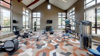 This fitness center is a thoughtfully designed workout space that combines striking architectural details with a comprehensive array of exercise equipment. The room commands attention with its soaring vaulted ceiling, accented by exposed wooden beams and clerestory windows that flood the space with natural light from multiple directions. Large floor-to-ceiling windows line the walls, offering open views of the surrounding landscape and lush greenery, creating an energizing and airy atmosphere throughout.

The flooring makes a bold design statement, featuring a vivid geometric pattern in terracotta, gray, and white tones that adds visual energy to the space. Cardio equipment, including treadmills and elliptical trainers, is positioned along the windowed walls to allow users to enjoy the outdoor views during their workouts. The strength-training area is well-stocked with selectorized cable machines, weight benches, and a full dumbbell rack, catering to a range of fitness goals.

Wall-mounted flat-screen televisions are distributed throughout the space, ensuring entertainment is always within view. A built-in shelving unit with a teal textured accent wall provides organized storage for accessories and equipment. The overall ambiance is modern and motivating, balancing functional fitness design with refined aesthetic touches that elevate the experience beyond a standard gym environment.