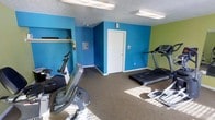 This single-floor fitness space offers a dedicated area for cardio-focused workouts in a compact yet functional layout. The room is immediately distinguished by its bold color scheme, with vivid blue walls dominating the space and a bright green accent wall adding an energizing contrast — a design choice well-suited to an active environment. The carpeted flooring provides a cushioned surface underfoot throughout the room. Equipment includes a treadmill and multiple stationary cardio machines, including a recumbent-style bike, giving users a variety of low-impact exercise options. Overhead fluorescent lighting ensures the space is well-lit, while a textured ceiling adds a classic finish. A built-in shelving nook along one wall offers convenient storage for personal items or accessories, and an electrical panel is accessible within the room. A white interior door provides access to an adjoining area. The overall ambiance is utilitarian and straightforward, with the vivid wall colors serving as the primary design element, creating an upbeat atmosphere that supports an active lifestyle. This fitness room is well-suited for residents or guests seeking a convenient space for daily cardio exercise.