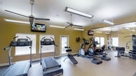 This fitness center is a dedicated single-floor exercise facility designed to support a well-rounded cardio workout routine. The room is finished with warm golden-yellow walls that create an energizing atmosphere, complemented by a light-colored ceiling that enhances the sense of openness. Natural light flows in generously through multiple white-trimmed windows fitted with blinds, as well as a classic French-style glass-panel door, keeping the space bright and inviting.

The facility is equipped with a thoughtfully arranged selection of cardio machines, including treadmills, elliptical trainers, and a stair stepper, along with a weight bench for strength training. Resistance bands are neatly stored on wall-mounted hooks, adding to the functional layout of the space. Two wall-mounted flat-screen televisions are positioned for easy viewing during workouts, providing entertainment to keep users engaged.

Overhead, multiple ceiling fans work in tandem with hanging linear light fixtures to ensure proper air circulation and consistent illumination throughout the room. The durable flooring is suited for high-traffic fitness use, offering both practicality and ease of maintenance. Overall, this fitness center delivers a comfortable, functional, and well-lit environment conducive to regular exercise.