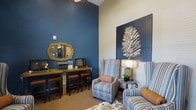 This single-floor space combines functionality and refined design to create a warm, inviting environment. The interior is anchored by a striking navy blue accent wall that rises to impressive heights, giving the space a sense of grandeur and depth. A decorative gold-framed mirror adds an elegant touch to the wall, complementing the rich, textured finishes throughout.

The business center area features a wood-toned counter with leather barstools, offering a practical workstation equipped with computer terminals — ideal for residents or visitors needing a dedicated workspace. Adjacent to this, a comfortable lounge area provides a relaxed setting with classic striped wingback chairs arranged around a stylish side table with a modern lamp. A large-scale pinecone artwork mounted on a dark panel serves as a bold focal point, reinforcing the space's nature-inspired aesthetic.

Warm carpeting and layered textural elements throughout contribute to an overall ambiance that is both cozy and sophisticated. The combination of deep navy tones, natural wood accents, and curated furnishings reflects a cohesive interior design vision that balances approachability with visual interest, making this space both practical and welcoming for everyday use.