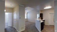 This single-story space showcases an open-concept layout that seamlessly connects the living area and kitchen. The living space is defined by vaulted ceilings that lend a sense of height and openness, complemented by a ceiling fan with a pendant light fixture that serves as a central focal point. Natural light filters in through multiple windows fitted with blinds, brightening the neutral gray and beige tones of the walls. The flooring in the living area consists of carpet, adding warmth underfoot, while hard surface flooring is present in the kitchen zone.

The kitchen features dark espresso-toned cabinetry that contrasts with a light-colored countertop extending into a breakfast bar, offering a casual dining or workspace option. A sink is integrated into the bar area, and stainless steel appliances are visible. A thermostat is mounted on a structural column near the entryway, which also features a classic six-panel white door. Floor vents are present throughout, indicating a central HVAC system. Overall, the space presents a clean, functional, and modern aesthetic with a comfortable, open feel.