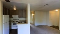 This single-floor space offers a practical and straightforward layout designed for comfortable everyday living. The kitchen is positioned in a semi-open configuration, separated from the main living area by a partial wall and countertop peninsula. It features a white gas range with overhead ventilation, a stainless steel sink, and dark wood-toned upper cabinetry that provides ample storage. The countertop extends to form a casual bar-style surface, making it convenient for meal prep and informal dining. The adjoining living area is finished with wall-to-wall carpet, lending a warm and quiet atmosphere to the space. Textured popcorn ceilings are consistent throughout, and ceiling-mounted light fixtures provide overhead illumination in the kitchen. A hallway visible from the main living area leads to additional rooms, suggesting a connected and navigable floor plan. The neutral, light-toned walls throughout keep the interior feeling open and easy to personalize. Overall, this space presents a no-frills, functional interior with a cohesive single-floor layout suitable for a variety of occupants.