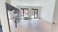 This contemporary single-floor space offers an open-concept layout that seamlessly integrates the kitchen and living area into one bright, cohesive environment. The kitchen is outfitted with dark cabinetry, stainless steel appliances including a refrigerator, stove, and built-in microwave, as well as a functional sink set beneath a window. The cabinetry's contrast against the white walls creates a clean, modern aesthetic throughout. Light-toned hardwood flooring extends across the entire space, contributing to an airy and welcoming atmosphere. A set of large, black-framed windows and a glass-paneled door line the exterior wall, inviting abundant natural light and offering views of the surrounding neighborhood. The windows are equipped with roller shades for added privacy and light control. Track lighting mounted on the ceiling provides flexible, even illumination throughout the open living area. The overall design is minimalist and functional, with a neutral color palette that provides a versatile backdrop well-suited to a variety of interior styles. The unit's thoughtful layout maximizes the sense of openness and flow from the kitchen through to the living space.