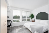 This thoughtfully designed single-floor studio presents a clean, modern aesthetic throughout its open-plan layout. Light wood-tone flooring and bright white walls create a fresh, airy atmosphere, while large windows along the main wall flood the space with natural light and frame views of the surrounding neighborhood and greenery.

The sleeping area occupies the central portion of the unit, complemented by an open-shelving wardrobe alcove near the entry, providing organized clothing and linen storage without occupying excessive floor space. A built-in workstation lines one wall, featuring a sleek gray desk surface, wall-mounted shelving above, and a full-height wardrobe cabinet alongside — a highly functional arrangement for studying or working from home.

The kitchenette is efficiently outfitted with light wood cabinetry, gray laminate countertops, a stainless steel sink, an electric cooktop, over-the-range microwave, and a full-size refrigerator. Open shelving beside the cabinetry offers additional storage flexibility.

Throughout the studio, the design prioritizes smart use of space with integrated built-ins that maximize functionality while maintaining an uncluttered, minimalist feel. Light-toned finishes, consistent flooring, and a neutral color palette contribute to a cohesive and inviting living environment.