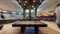 This expansive amenity lounge is defined by its dramatic double-height ceilings and an industrial-chic design that blends raw materials with refined finishes. The centerpiece of the main gathering area is a full-size billiards table set atop wide-plank hardwood flooring, surrounded by an open layout that encourages socializing. Overhead, a striking ceiling clad in mixed-metal tiles and globe pendant lights adds visual depth and character.

A dedicated lounge zone along one wall features an extended banquette with upholstered seating, decorative stone accents, and an eye-catching black pipe-work wall installation fitted with Edison-style sconces. This area is complemented by eclectic armchairs and dark marble-topped tables, creating an intimate atmosphere within the larger open space.

A generously sized bar and kitchen island anchors the hospitality area, showcasing a bold granite countertop, bar-height seating, pendant lighting, and exposed brick walls with rustic wood shelving. A large wall-mounted display screen and dark cabinetry complete the setup.

Floor-to-ceiling steel-framed glass windows and doors flood the space with natural light and provide views of an outdoor courtyard. Adjacent to the main lounge, a glass-enclosed fitness center is visible through black-framed partitions, maintaining visual openness throughout.