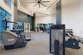 Located at 925 W 20th St in Houston's vibrant Heights neighborhood, this fitness center offers a spacious, single-floor layout designed to accommodate a full range of workout routines. The space is immediately striking thanks to its dramatically high ceilings, which lend an open, airy atmosphere throughout. A large statement ceiling fan anchors the room overhead, complemented by sleek linear pendant lighting that enhances the modern aesthetic.

The interior design features a bold yet refined color palette, with deep teal accent walls and a standout decorative mural depicting oversized golden keys — adding a distinctive artistic touch to the workout environment. Large floor-to-ceiling windows along one wall flood the space with natural light and provide views to the surrounding exterior.

The fitness floor is comprehensively outfitted for both cardio and strength training. Cardio equipment includes treadmills, stair climbers, stationary bikes, and rowing machines, while the strength training area features cable machines, a functional training rig, free weights, kettlebells, medicine balls, battle ropes, and a full rack of dumbbells. Floor-mounted mirrors expand the sense of space in the strength zone, and a wall-mounted TV keeps members engaged during workouts.

A built-in storage and refreshment station with teal cabinetry rounds out the space, providing a convenient amenity area. The durable tile-pattern carpet flooring ties the entire room together for a polished, professional finish.