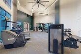 Located at 925 W 20th St in Houston's vibrant Heights neighborhood, this fitness center offers a spacious, single-floor layout designed to accommodate a full range of workout routines. The space is immediately striking thanks to its dramatically high ceilings, which lend an open, airy atmosphere throughout. A large statement ceiling fan anchors the room overhead, complemented by sleek linear pendant lighting that enhances the modern aesthetic.

The interior design features a bold yet refined color palette, with deep teal accent walls and a standout decorative mural depicting oversized golden keys — adding a distinctive artistic touch to the workout environment. Large floor-to-ceiling windows along one wall flood the space with natural light and provide views to the surrounding exterior.

The fitness floor is comprehensively outfitted for both cardio and strength training. Cardio equipment includes treadmills, stair climbers, stationary bikes, and rowing machines, while the strength training area features cable machines, a functional training rig, free weights, kettlebells, medicine balls, battle ropes, and a full rack of dumbbells. Floor-mounted mirrors expand the sense of space in the strength zone, and a wall-mounted TV keeps members engaged during workouts.

A built-in storage and refreshment station with teal cabinetry rounds out the space, providing a convenient amenity area. The durable tile-pattern carpet flooring ties the entire room together for a polished, professional finish.