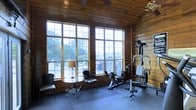 This inviting single-floor space showcases a distinctive rustic character defined by warm, honey-toned horizontal wood-paneled walls and a matching wood-planked ceiling that together create a cozy, lodge-like atmosphere. Large, multi-pane windows dominate one wall, flooding the interior with generous natural light and framing lush outdoor views of surrounding greenery and what appears to be a deck or patio area beyond. A ceiling fan overhead ensures comfortable air circulation throughout. The space thoughtfully combines two distinct functions: a dedicated fitness area equipped with a multi-station home gym machine and exercise equipment, complete with protective rubber flooring tiles, and a relaxed lounge corner furnished with sleek black armchairs and a glass-topped accent table with a classic table lamp. This dual-purpose layout makes the room equally suited for active workouts and quiet relaxation. The overall ambiance strikes a balance between rustic warmth and functional design, making it a versatile and welcoming environment year-round.