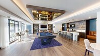 Located at 37 N York Rd in Hatboro, Pennsylvania, this single-floor space seamlessly blends a fully appointed kitchen with an open-concept living area, offering a cohesive and thoughtfully designed environment throughout.

The kitchen is a standout feature, anchored by an expansive white quartz island with seating for multiple bar-height chairs finished in gold-toned metal frames. Elegant globe pendant lights with brass hardware hang above the island, casting a warm glow over the space. Dark wood cabinetry lines the perimeter walls, complemented by blue mosaic tile backsplash and open floating shelves. A dedicated counter area provides workspace and appliance storage, including a coffee station and sink.

The living area flows naturally from the kitchen and is centered around a built-in entertainment wall with dark wood shelving, a large wall-mounted television, and a sleek marble-surround fireplace. A generous leather sofa anchors the seating area, accented by a blue area rug and modern side tables. Recessed tray ceilings with ambient lighting add architectural depth and a refined finish to the room.

Floor-to-ceiling windows along the exterior wall flood the space with natural light and offer views to the outdoors, where bistro-style high-top tables and chairs create a connected lounge area. Warm hardwood flooring runs throughout, tying together the kitchen and living zones into one inviting and functional space.