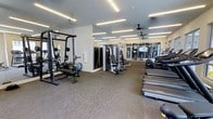 This fitness center offers a thoughtfully designed workout environment that balances functionality with a clean, contemporary aesthetic. The single-floor layout provides an open, spacious feel, with neutral-toned carpet flooring throughout that helps define workout zones while reducing noise. Linear LED lighting runs across the ceiling, ensuring consistent, bright illumination across the entire space.

The strength training area features a multi-function cable machine, a smith machine with an attached bench, and a variety of free weights and accessories, catering to a wide range of resistance-based exercises. The cardio section, positioned toward the rear of the space, houses multiple treadmills and additional cardio equipment lined along the perimeter, allowing users to work out while enjoying views through large glass doors and windows that bring in natural light.

Ceiling fans are strategically placed to promote airflow and comfort during intense workouts. Mirrored wall panels enhance the sense of depth and openness while serving a practical purpose for form-checking. The overall ambiance is modern and inviting, striking a balance between a professional gym setting and an accessible community fitness facility.