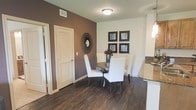 This single-floor residence combines contemporary style with warm, inviting design elements throughout. The dining area serves as a standout feature, showcasing rich, dark hardwood flooring that grounds the space with a sense of elegance. A bold mocha-toned accent wall creates a striking focal point, complemented by a decorative sunburst mirror and a curated gallery of framed black-and-white artwork, adding depth and visual interest to the room. The dining space accommodates a round wooden table surrounded by crisp white upholstered chairs, offering both functionality and sophistication. A louvered closet door provides practical storage while contributing to the clean, classic aesthetic of the space. Adjacent to the dining area, a glimpse of the kitchen reveals granite countertops in neutral tones, seamlessly connecting the living and dining areas. Flush-mount ceiling lighting provides consistent illumination throughout. The overall ambiance is polished and cohesive, with a carefully balanced contrast between the dark flooring, white walls, and warm accent tones — creating a space that feels both modern and comfortable.