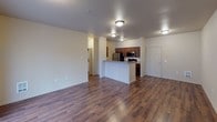 Located at 10436 SE Carr Rd in Renton, Washington, Unit A401 is a thoughtfully designed single-floor residence that offers a comfortable and functional layout. The open-concept living and dining area is finished with rich, dark hardwood-style flooring that extends throughout the main living spaces, creating a warm and cohesive aesthetic. A sliding glass door in the living area allows natural light to flow in and provides access to an outdoor space. The kitchen is positioned centrally and features a peninsula-style breakfast bar, rich wood cabinetry, a stainless steel refrigerator, and black appliances including a range and microwave. The entry foyer connects directly to the kitchen corridor, offering a practical and efficient flow upon arrival. The bedrooms are carpeted for added comfort and warmth, each offering generous proportions. One bedroom features a large window that brings in natural light, while another includes a sliding closet with a rod and shelf for organized storage. The bathroom is well-equipped with a single-sink vanity with wood cabinetry, a full tub and shower combination, a large mirror, and tile flooring. Neutral wall tones throughout the unit lend a clean, versatile backdrop suitable for a variety of interior styles.