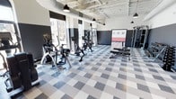 This single-floor fitness center is designed with both functionality and aesthetics in mind. The open layout provides ample space for a diverse range of workout equipment, including elliptical machines, stationary bikes, and a multi-use cable and weight training station. A weight bench and free weights are also available, catering to a variety of fitness routines.

The space features an eye-catching checkered floor in shades of gray and white, adding a dynamic visual element to the room. The walls are painted in a bold dark charcoal with contrasting white upper sections, giving the gym a contemporary, energetic atmosphere. An accent wall with a red and white panel adds a striking focal point to the space.

The exposed industrial ceiling showcases structural beams and ductwork, complemented by sleek black pendant lights that provide focused overhead lighting. Large floor-to-ceiling windows along one wall flood the interior with natural light, offering views of the surrounding streetscape and creating an open, inviting environment. The combination of modern equipment, thoughtful design elements, and abundant natural light makes this fitness center a well-rounded and motivating space for exercise.