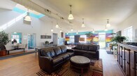 This single-floor community clubhouse is a lively, well-appointed gathering space designed for recreation and socializing. The main area is expansive and open, featuring soaring vaulted ceilings with exposed ductwork that lend an industrial character, complemented by rows of pendant lights casting warm, even illumination across the hardwood floors throughout.

The lounge zone anchors one end of the space with dark leather sofas, an ottoman, and a patterned area rug, creating a comfortable seating arrangement. Adjacent to it, a full-size Olhausen pool table serves as a centerpiece for the game area, accompanied by a ping-pong table and an oversized Connect Four set. High-top seating near large windows and a glass exterior door allows natural light to pour in, offering views of surrounding greenery.

A boldly painted tropical sunset mural along the teal-accented walls adds vivid color and personality throughout. A glass-enclosed meeting or dining room provides a quieter, separated space within the layout.

Additional amenities include a dedicated tanning room equipped with a stand-up tanning bed and a wooden bench, and a compact kitchenette outfitted with warm wood cabinetry, dark countertops, a stainless steel sink, and a microwave. Together, these features create a diverse, amenity-rich environment suited for both relaxation and active recreation.