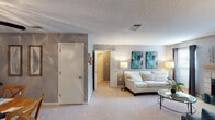 This single-floor residence offers a thoughtfully designed layout across multiple rooms, each finished in a consistent palette of soft grey and neutral tones with plush wall-to-wall carpeting throughout the living areas. The living room serves as a central gathering space, highlighted by a tiled wood-burning fireplace with a wood mantle, large windows dressed in dark curtain panels, and an open flow into an adjacent sitting or flex room outfitted with teal accent chairs and natural light. The galley-style kitchen features warm wood cabinetry, light countertops, tile flooring, and a full suite of appliances, opening directly into the carpeted dining area, which is anchored by a ceiling fan and comfortable seating for four. The primary bedroom is generously proportioned, complete with a walk-in closet and direct access to a full bathroom. A secondary bedroom accommodates twin beds and benefits from natural light through a window with curtain panels. The bathrooms are finished cleanly with neutral countertops, a full vanity mirror, and tub-shower combinations. Textured ceilings are present throughout, and the overall ambiance is calm, cohesive, and move-in ready.