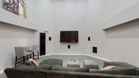 This single-floor space centers around a thoughtfully designed media lounge that prioritizes comfort and entertainment. The room immediately draws attention with its dramatically high ceilings, which lend an expansive, open feel to the interior. Neutral light gray walls create a clean, modern backdrop that complements the space's minimalist aesthetic.

A large wall-mounted flat-screen television serves as the focal point, flanked by built-in in-wall speakers positioned strategically throughout the room to deliver a full surround sound experience. Additional speakers are mounted at varying heights along the walls, reinforcing the space's dedication to high-quality audio performance.

The seating arrangement features a generous sectional sofa in a muted gray fabric, accompanied by a central ottoman that doubles as a coffee table. Accent pillows in warm tones provide a subtle contrast to the otherwise cool palette. Soft carpeting underfoot adds a layer of comfort suited to extended viewing sessions.

A black-framed door on the left side of the room provides access to an adjacent area, while a small seating nook is partially visible beyond it, suggesting additional functional space nearby. Overall, the room balances modern design with practical amenities.